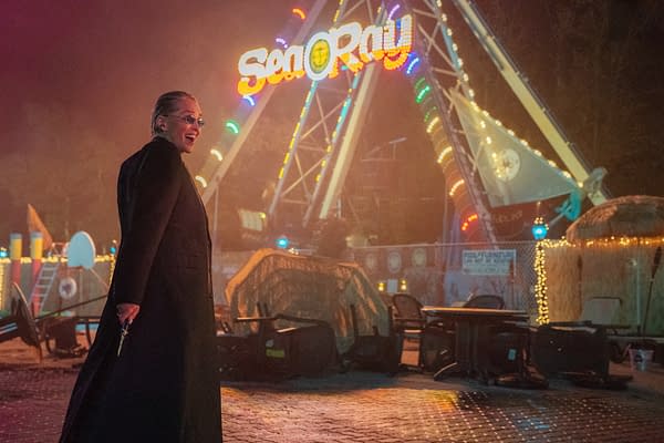 A woman in a long black coat stands in front of a brightly lit amusement ride named 'Sea Ray' at night, exuding confidence as she holds a small object in her hand. The scene is illuminated with colorful lights, creating an atmospheric and slightly mysterious vibe.