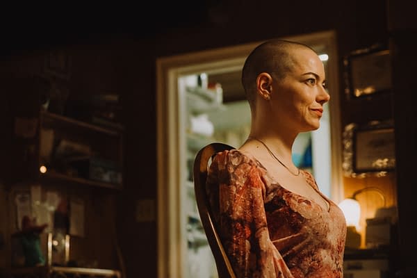 Emma Stone portrays a character named Michelle in the film 'BUGONIA', directed by Yorgos Lanthimos. The image features her with a shaved head and a floral dress, set against a backdrop that suggests a cozy interior.