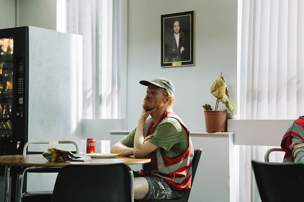 A scene from the film 'BUGONIA' featuring Jesse Plemons as Teddy Gatz, seated at a table in a cafeteria, wearing a green shirt and red-striped vest, with a can of Coca-Cola and snacks nearby. In the background, there is a vending machine and a framed portrait on the wall.