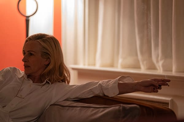 A woman with long, blonde hair sits on a couch, dressed in a white shirt, casually resting her arm with a cigarette. The setting features soft lighting and a warm orange wall, contributing to a contemplative atmosphere.