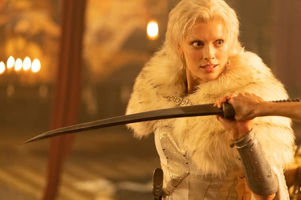 A scene from the new Red Sonja movie featuring a character with long blonde hair and a fur cloak, engaged in a tense moment while holding a sword. The setting is dimly lit, emphasizing the character's focused expression.