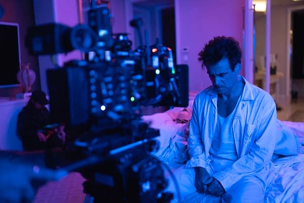 A scene from 'Ballad of a Small Player' showing a male actor in a white outfit, sitting on a bed with a somber expression, while a film camera is focused on him. The room is illuminated with blue and purple lighting, suggesting an emotional or dramatic moment.