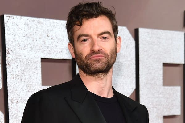 A man with medium-length wavy hair and a beard poses confidently in a black suit against a textured background with large, white letters. He is featured in a post about Zack Snyder's upcoming film, 'The Last Photograph.'