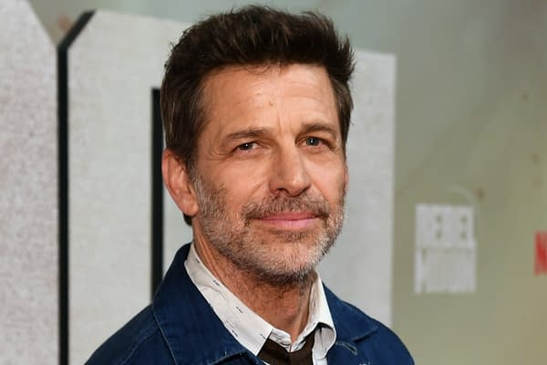 Zack Snyder smiling at a premiere event, wearing a denim jacket over a collared shirt, with a blurred background featuring promotional elements.