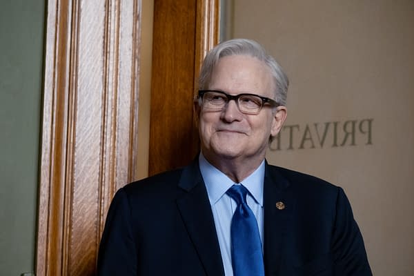 A scene from the film 'ELLA MCCAY' featuring Albert Brooks portraying Governor Bill, dressed in a dark suit and light blue shirt with a matching tie, smiling in a warmly lit interior.