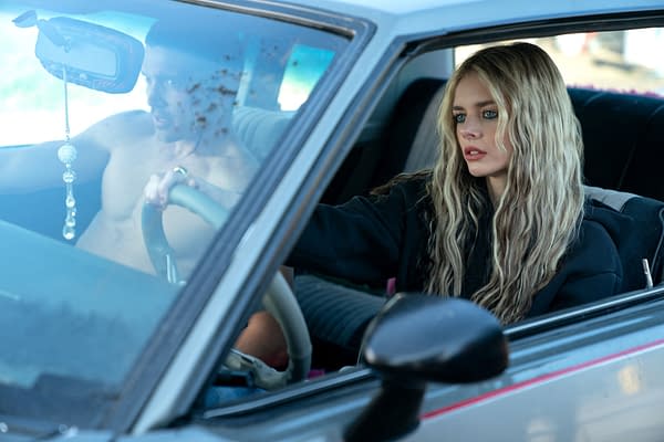 A scene from 'Eenie Meanie' featuring Karl Glusman as John driving a car while shirtless, with Samara Weaving as Edie sitting beside him. The interior of the car shows a blurred view of a rearview mirror and dashboard decorations.
