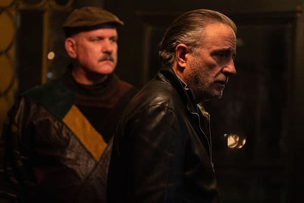 A scene from the film 'Eenie Meanie' featuring Mike O'Malley as George in a colorful, patterned sweater and Andy Garcia as Nico in a black leather jacket, both in a dimly lit environment, conveying a tense moment.