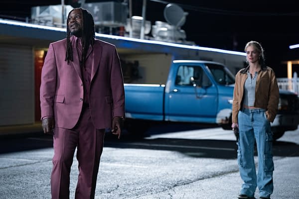 A scene from 'EENIE MEANIE' featuring Marshawn Lynch dressed in a pink suit with long hair and Samara Weaving in casual attire, standing in front of a blue pickup truck at night.