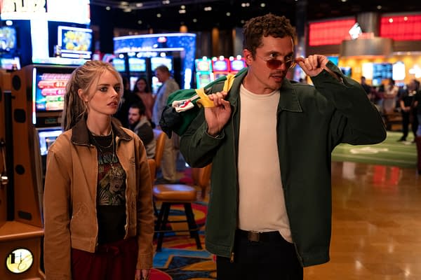 Still from the movie 'Eenie Meanie' featuring characters Edie, portrayed by Samara Weaving, and John, played by Karl Glusman, in a vibrant casino setting with slot machines in the background.