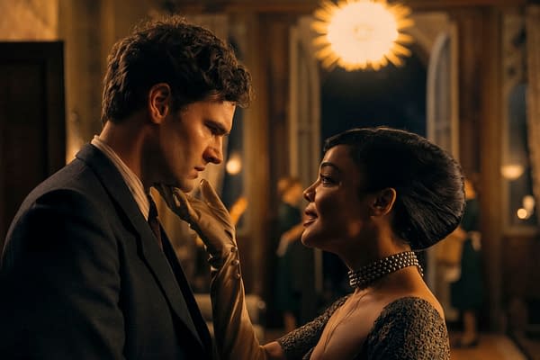 A scene from 'HEDDA' featuring Tessa Thompson as Hedda Gabler and Tom Bateman as George Tessman, sharing an intimate moment. The setting is a dimly lit room with a stylish chandelier in the background, emphasizing the dramatic tone of the play.