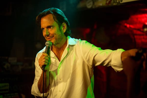 Will Arnett performing stand-up comedy with a microphone in hand, wearing a white shirt, under colorful stage lighting. The setting appears to be an intimate venue.