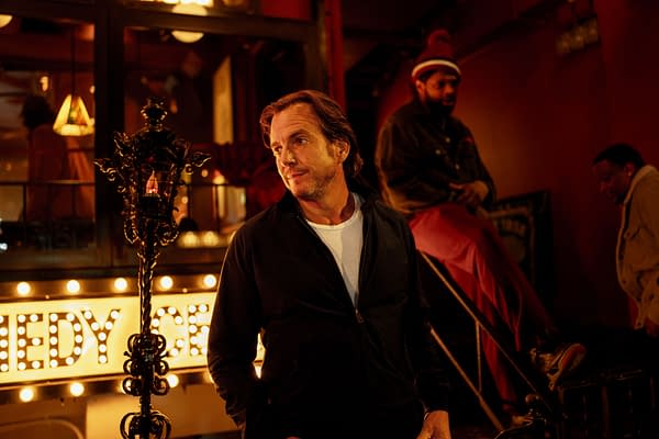 Will Arnett stands in front of a brightly lit comedy club sign, exuding a relaxed demeanor in a dimly lit setting, while two other individuals are seen in the background.