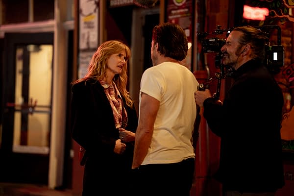 Laura Dern and Will Arnett are seen engaged in conversation on the set of the film 'IS THIS THING ON?' with director Bradley Cooper also present. The scene is set on a vibrant street, illuminated by warm lights.