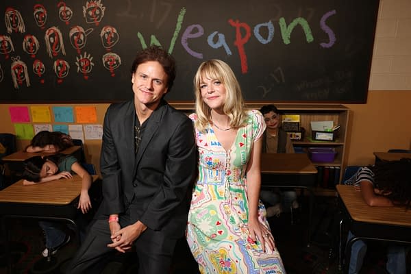 A promotional illustration featuring two actors, one wearing a black suit and the other in a colorful patterned dress, seated in a classroom setting. Behind them is a chalkboard with the word 'Weapons' written in colorful letters, with students visible in the background appearing to rest their heads on their desks.