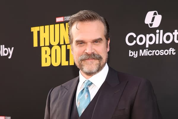 David Harbour smiles at the premiere of 'Thunderbolts,' dressed in a dark suit with a light blue tie, set against a black backdrop featuring the movie title.