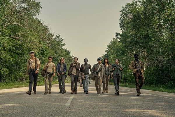 A group of nine young men walks together along a deserted road surrounded by lush greenery, each carrying bags and cameras, likely depicting a scene from the film 'The Long Walk.' The characters feature diverse clothing styles and expressions, highlighting a sense of camaraderie.