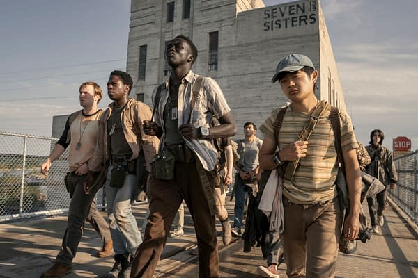 A group of young men, dressed in casual clothing with bags and accessories, walk together along a pathway near an industrial building. The scene captures a sense of camaraderie and determination as they head towards an unknown destination.