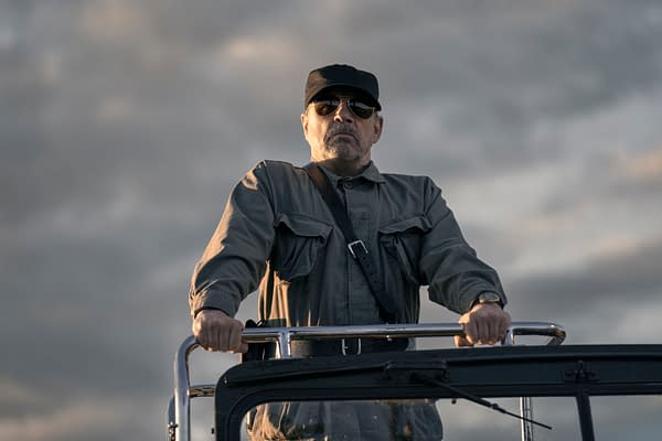 A man wearing sunglasses and a cap stands at the helm of a vehicle, dressed in a military-style shirt with pockets. He appears serious and commanding against a cloudy sky backdrop, portraying the character The Major in 'The Long Walk'.