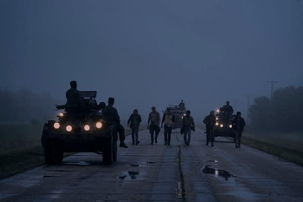 A scene from 'The Long Walk' featuring a group of individuals walking on a foggy road, flanked by military vehicles. The atmosphere is tense and dramatic, highlighting the journey of the characters.