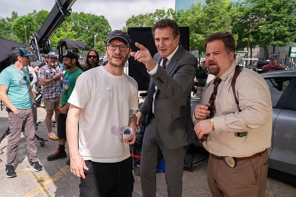 On the set of 'The Naked Gun,' director Akiva Schaffer interacts with actors Liam Neeson and Paul Walter Hauser, while crew members are seen in the background. The scene captures a moment of collaboration in a bustling film environment.