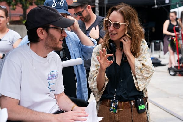 On the set of The Naked Gun, director Akiva Schaffer and producer Erica Huggins engage in conversation, with Huggins gesturing animatedly. The background includes crew members and equipment typical of a film shoot.