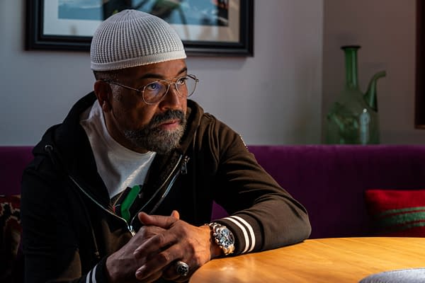 A thoughtful man with a beard and glasses, wearing a white knit cap and a black jacket, sits at a wooden table. The background features a purple couch and a green glass object, with soft lighting that highlights his introspective expression.