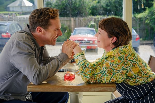 A cozy scene from 'The Roses' featuring two characters at a table, engaged in a warm conversation while holding hands. One person wears a colorful patterned shirt and the other a casual grey shirt, with a glass of red drink visible on the table.