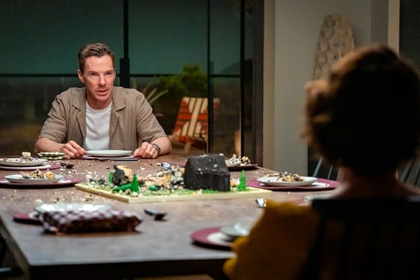 A scene from the film 'The Roses' featuring a man, seated at a dining table with a disheveled meal setup, expressing tension. The table is adorned with various plates and an elaborate centerpiece resembling a house made of food.
