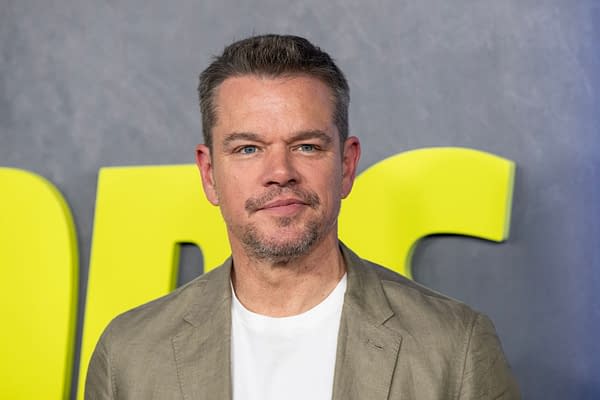 Actor Matt Damon poses at the premiere of 'The Instigators' in New York, wearing a light blazer and white t-shirt, with a bold yellow background.