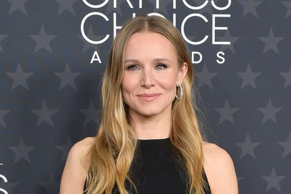 Kristen Bell smiles at an awards event, wearing a black dress with long, wavy hair, in front of a star-patterned backdrop. This image is associated with the announcement of her joining the cast of 'Violent Night 2'.