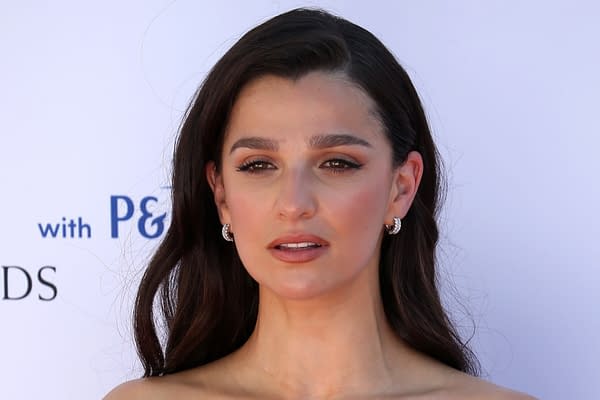 A close-up portrait of a woman with long dark hair, wearing elegant earrings and a natural makeup look, set against a light background featuring logos.