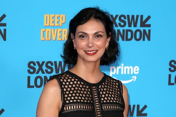 A woman with short, dark hair smiles confidently in front of a bright blue background featuring the text 'SXSW LONDON' and 'DEEP COVER.' She wears a stylish black dress with intricate cutouts, exuding elegance at a promotional event.
