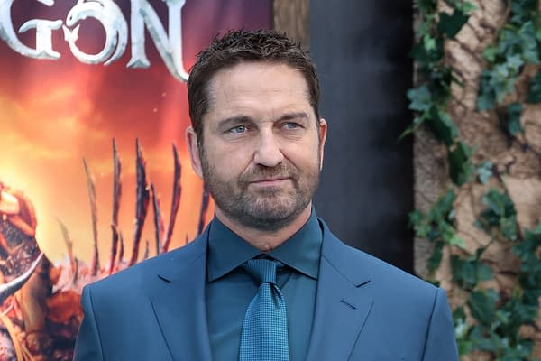 A man in a formal blue suit and tie stands in front of a colorful promotional backdrop featuring a fantasy theme. His expression is serious, and he has short, neatly styled hair and a beard.