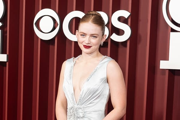 A woman with red hair and bold red lipstick smiles while wearing a stunning silver gown adorned with embellishments. Behind her is a backdrop featuring the CBS logo.