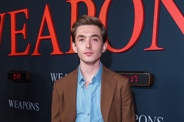 A young man stands in front of a dark backdrop with the word 'WEAPONS' in bold orange letters. He is wearing a brown blazer over a light blue shirt, looking directly at the camera.