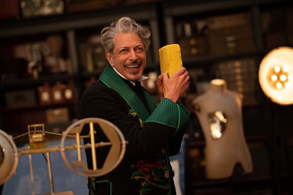In WICKED FOR GOOD, Jeff Goldblum, portraying The Wizard of Oz, enthusiastically holds a large yellow object while wearing an ornate black coat with green accents amidst a backdrop of vintage items.