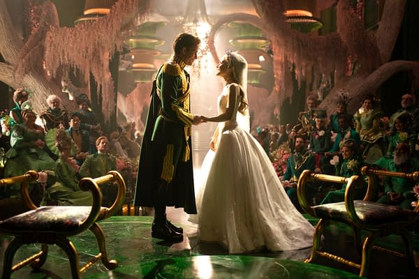 In a scene from 'WICKED FOR GOOD,' actors Jonathan Bailey and Ariana Grande, as Fiyero and Glinda, respectively, hold hands in an opulent setting adorned with greenery and flowers, while an audience in vibrant costumes watches.