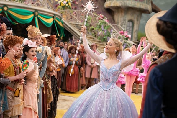 Ariana Grande, dressed as Glinda in a sparkling gown and crown, joyfully raises a wand in a whimsical scene filled with colorful characters and a festive atmosphere. The setting appears to be from the musical 'WICKED FOR GOOD', directed by Jon M. Chu.