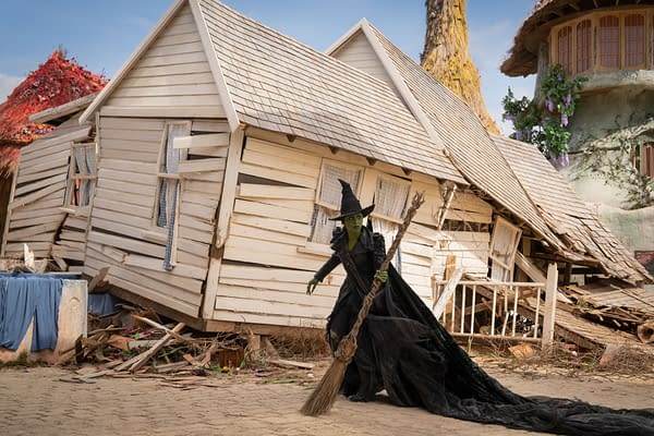 Cynthia Erivo portrays Elphaba in a scene from the film 'WICKED FOR GOOD,' featuring her wearing a dark, flowing gown and wielding a broomstick in front of a tilted, damaged house.