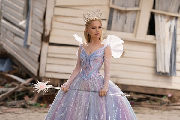 Ariana Grande as Glinda, dressed in an elaborate, shimmering gown with a silver tiara, holding a sparkling wand, set against a backdrop of a weathered house.