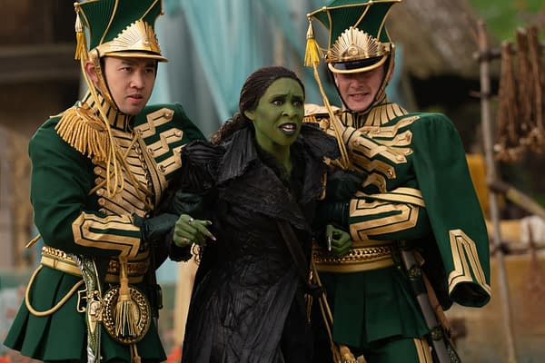 Cynthia Erivo portrays Elphaba in 'WICKED FOR GOOD', surrounded by two men in ornate green and gold costumes, depicting characters from the musical. The scene captures a dramatic moment with Elphaba looking concerned as she is restrained by the guards.