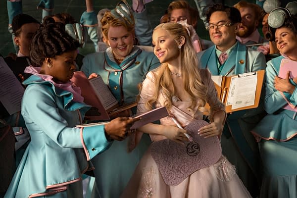A scene from 'Wicked: For Good' featuring Ariana Grande as Glinda, surrounded by a group of elegantly dressed characters in turquoise outfits. They appear to be in a joyful and engaging moment, holding music sheets and smiling.