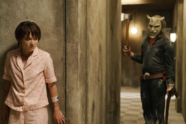 A scene from 'Black Phone 2' featuring a young girl in pink pajamas looking frightened against a concrete wall as a masked figure holding a weapon looms in the background.