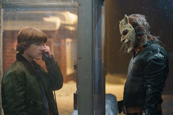 A tense confrontation occurs between a young man speaking on a payphone and a figure wearing a grotesque mask, set against a snowy backdrop, hinting at a suspenseful scene.