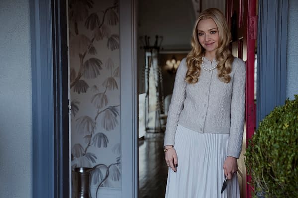 A young woman with long, wavy blonde hair stands by a doorway, wearing a light gray, cable-knit sweater and a flowing white skirt. She smiles warmly, holding a small object in her hand, with a stylish interior visible behind her.