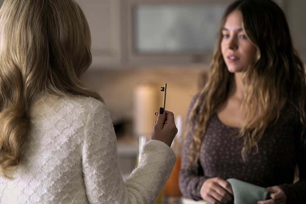 A scene from 'The Housemaid' featuring two characters: one woman, dressed in a white outfit with wavy hair, holds a gold key, while another woman with long, wavy hair, dressed in a brown patterned top, looks at her with intrigue. The setting appears to be a kitchen.