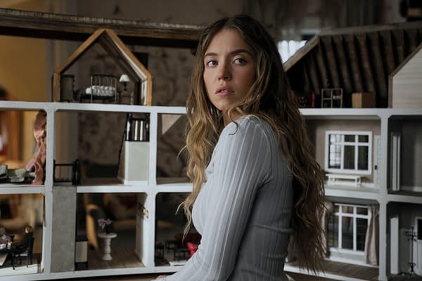 A young woman with long, wavy hair is sitting beside a detailed dollhouse, looking thoughtfully to the side. She appears to be wearing a fitted white top, enhancing a sense of contemplative emotion.