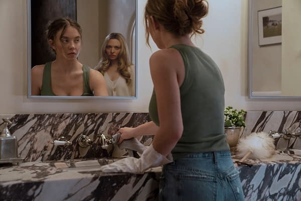 A scene from 'The Housemaid' showing a woman cleaning a sink while another woman appears in the background, watching her through a mirror. The contrasting expressions suggest tension between the characters.