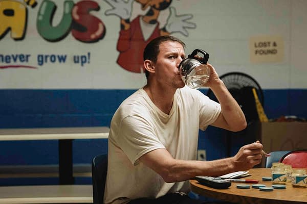 Channing Tatum sits at a table, drinking from a glass pitcher while holding a spoon, in a setting with a colorful wall that reads 'Toys R Us.' The scene conveys a casual, reflective moment.