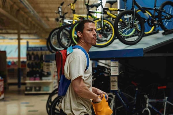 In a scene from Paramount Pictures' "ROOFMAN," Channing Tatum stands in a sporting goods store, holding an orange bag, while wearing a colorful backpack and looking contemplatively at the bicycles displayed in the background.